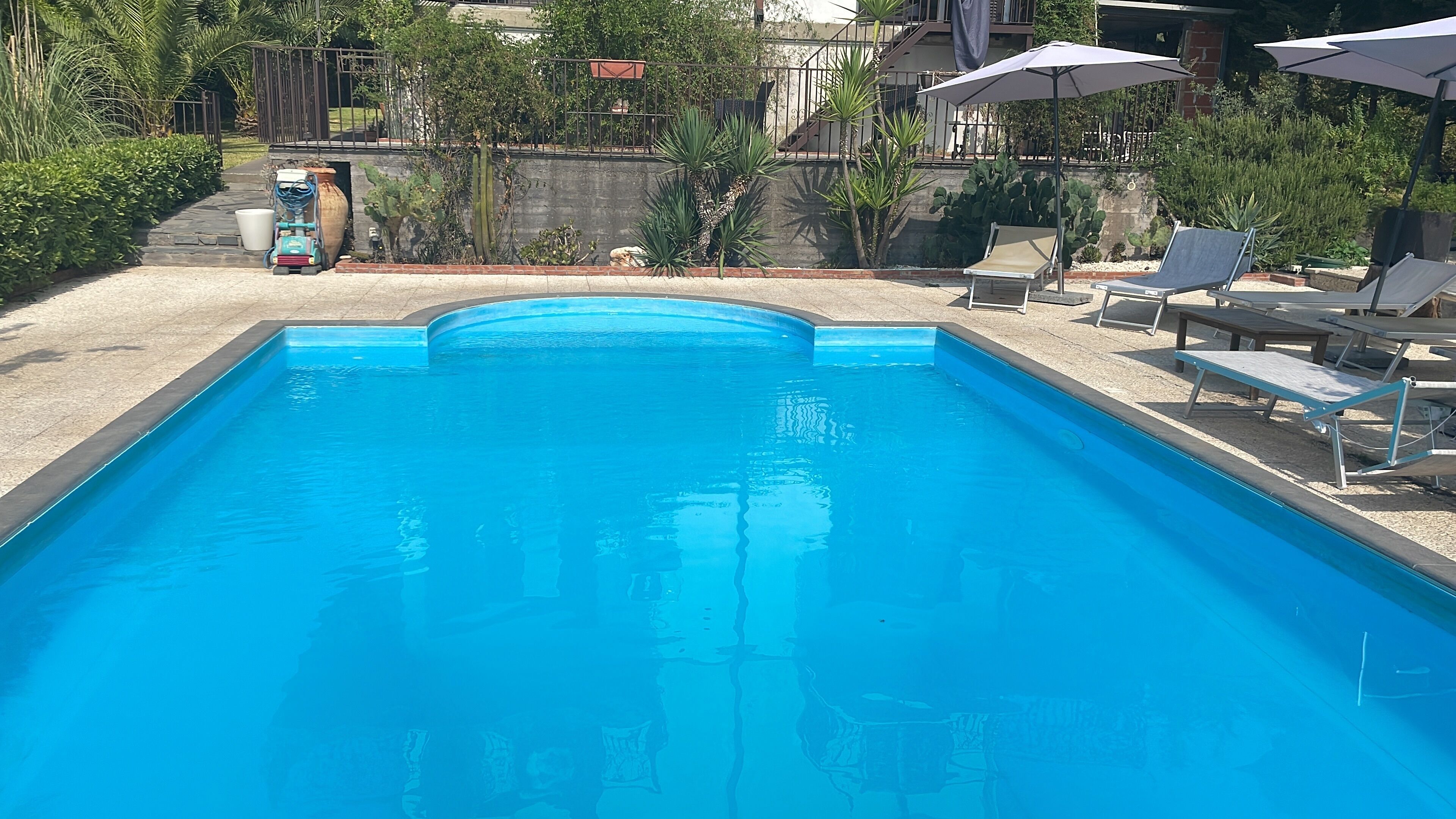 Una piscina rectangular con agua azul cristalina, rodeada por una solana de hormigón. Hay sillones de descanso y sombrillas en el lado derecho.