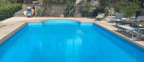 Una piscina rectangular con agua azul cristalina, rodeada por una solana de hormigón. Hay sillones de descanso y sombrillas en el lado derecho.