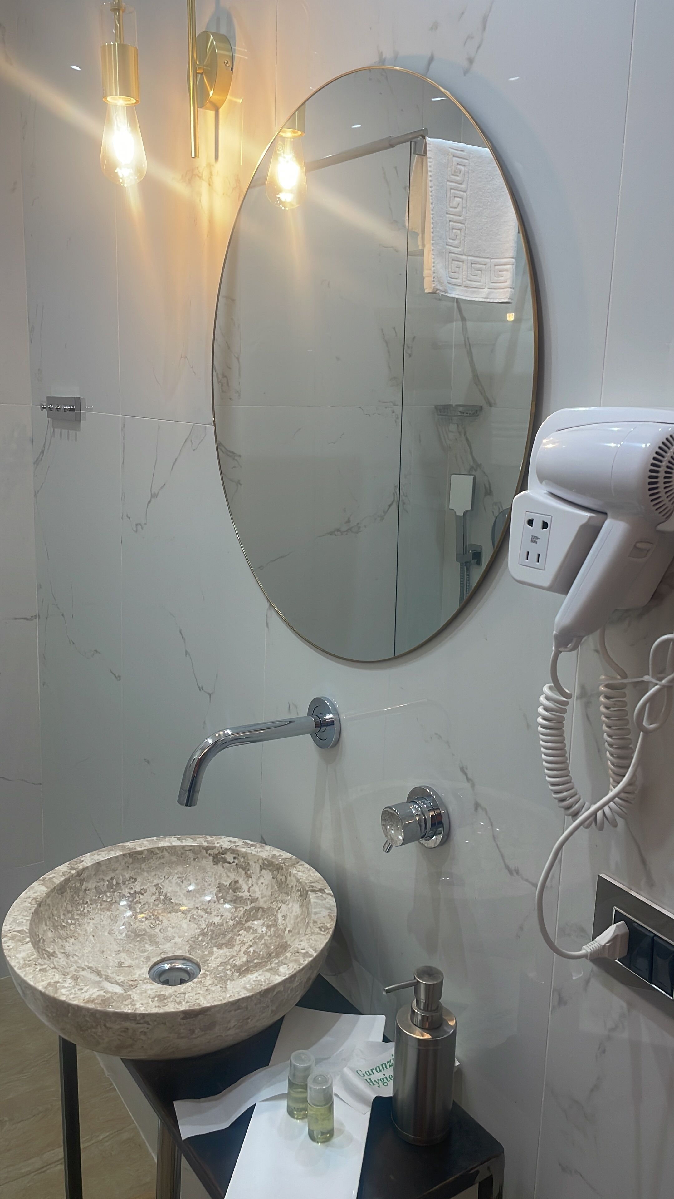 Une salle de bain avec un lavabo en marbre, un miroir rond, un sèche-cheveux et deux lampes fixées au mur.