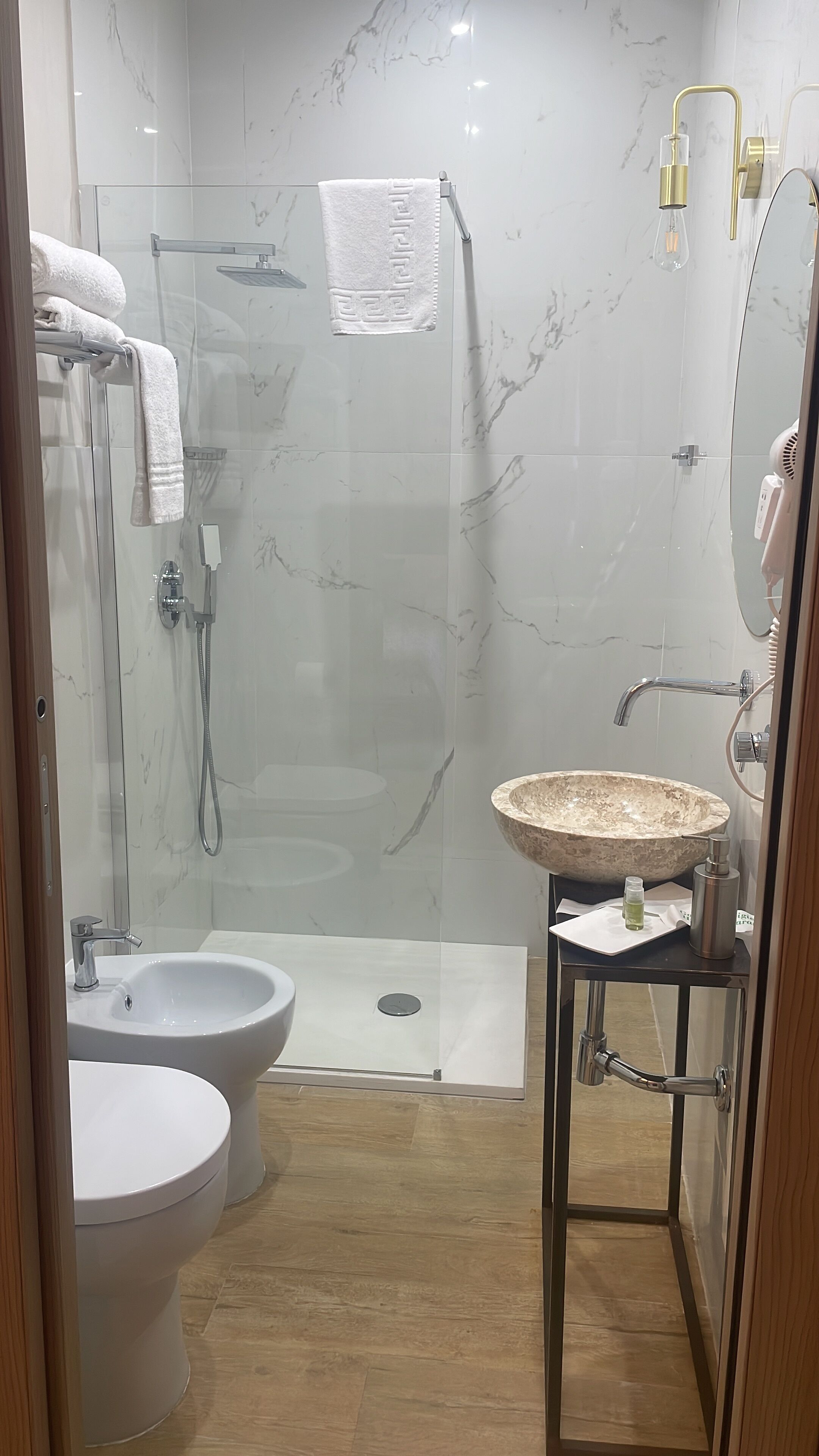 A modern bathroom with a glass-enclosed shower, two white basins, a marble-tiled wall, and a wooden floor.