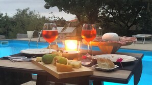 A poolside table with a fruit platter, two glasses of red wine, and a bowl of bread and cheese.