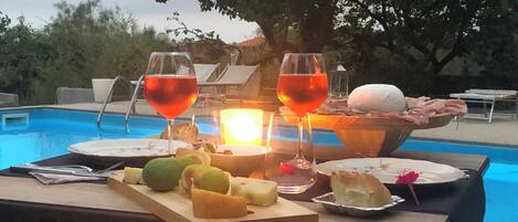 Une table placée au bord de la piscine, avec un plateau de fruits, deux verres de vin rouge et un panier de pain et de fromage.