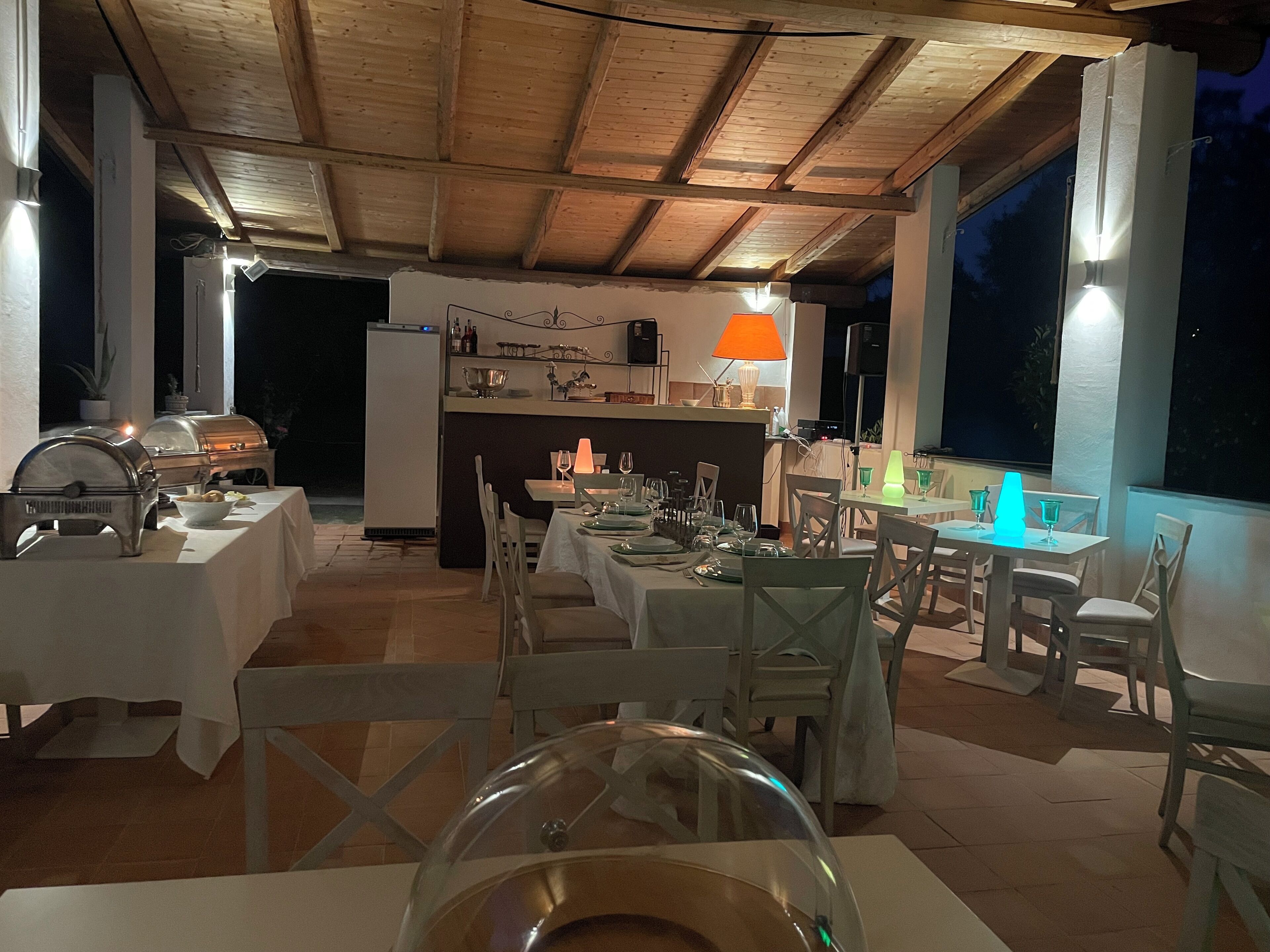 An outdoor dining area with tables set for a meal, a grill, and a kitchen area in the background.