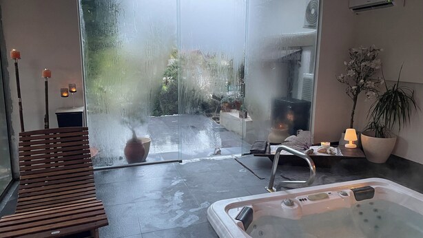 A modern bathroom with a glass shower, a wooden bench, a hot tub, and a potted plant.