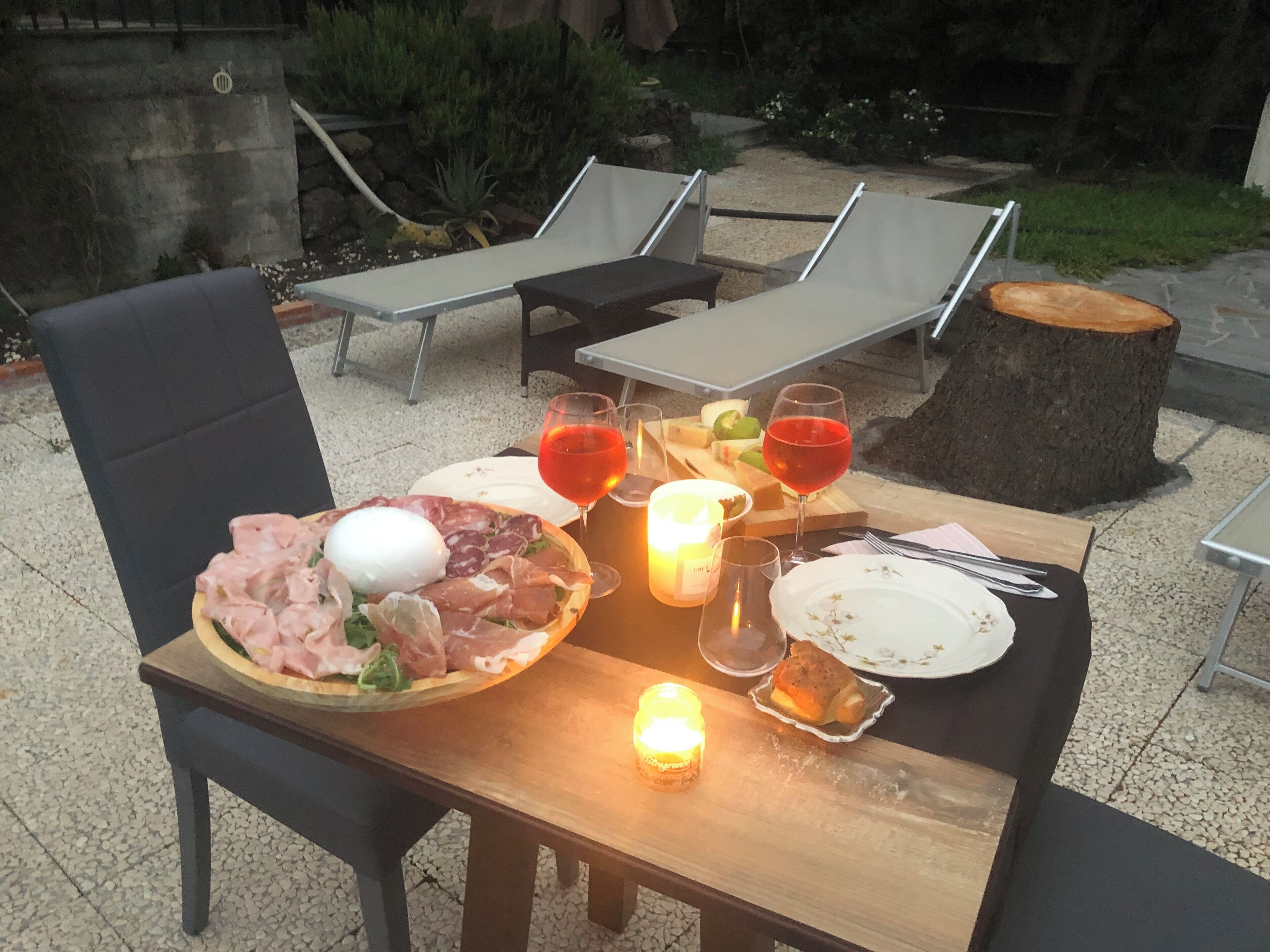An outdoor dining setup with a table set for two, featuring a variety of foods and drinks, surrounded by lounge chairs and a garden view.