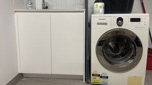 Een compacte, witte wasmachine met een digitaal scherm en een voorlaadsluiting, geplaatst in een kleine wasruimte met een betegelde spatwand en een witte wasbak.