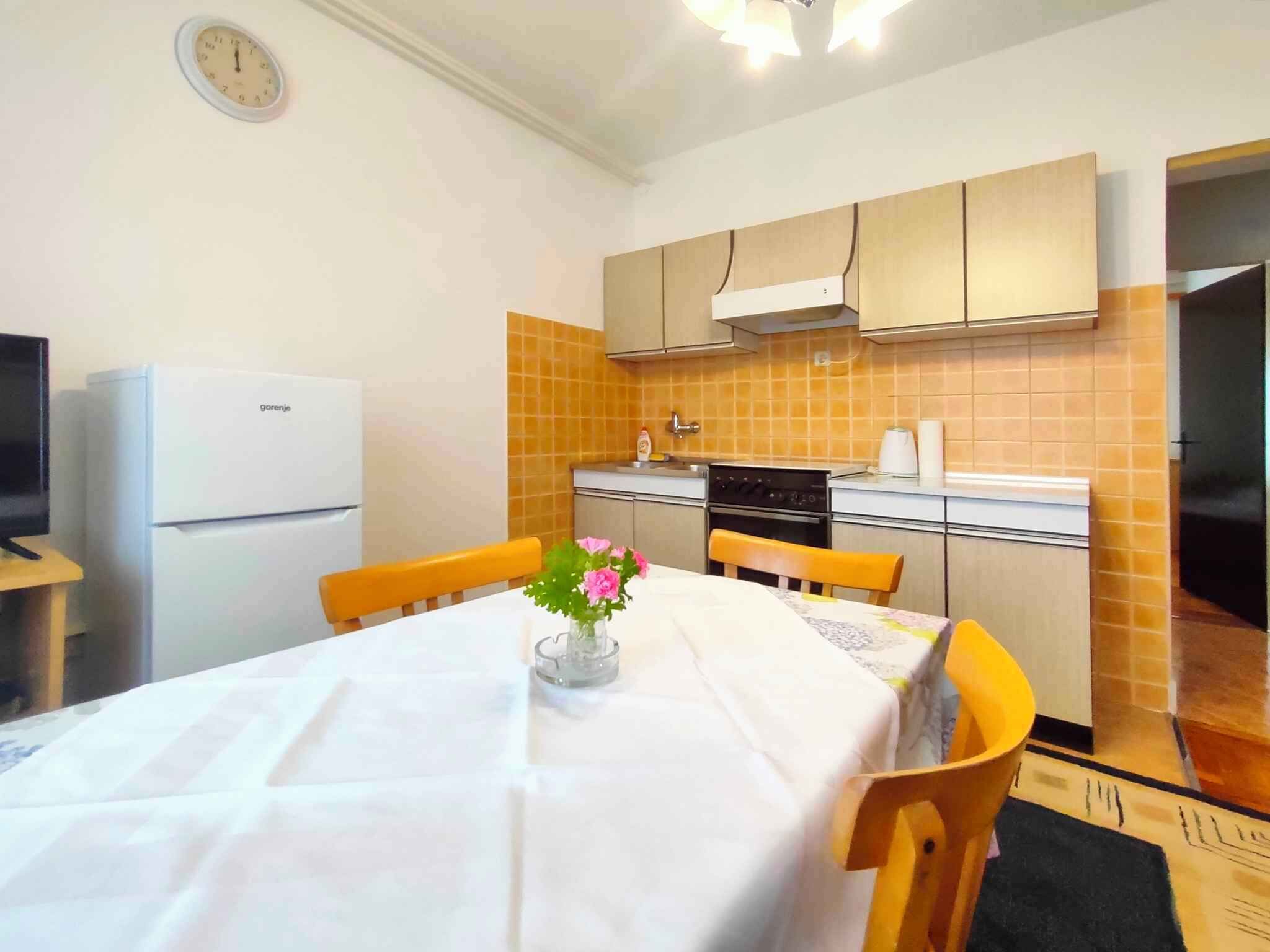 A compact kitchen with a refrigerator, stove, and orange-tiled backsplash. A dining table with chairs and a vase of flowers is in the foreground.