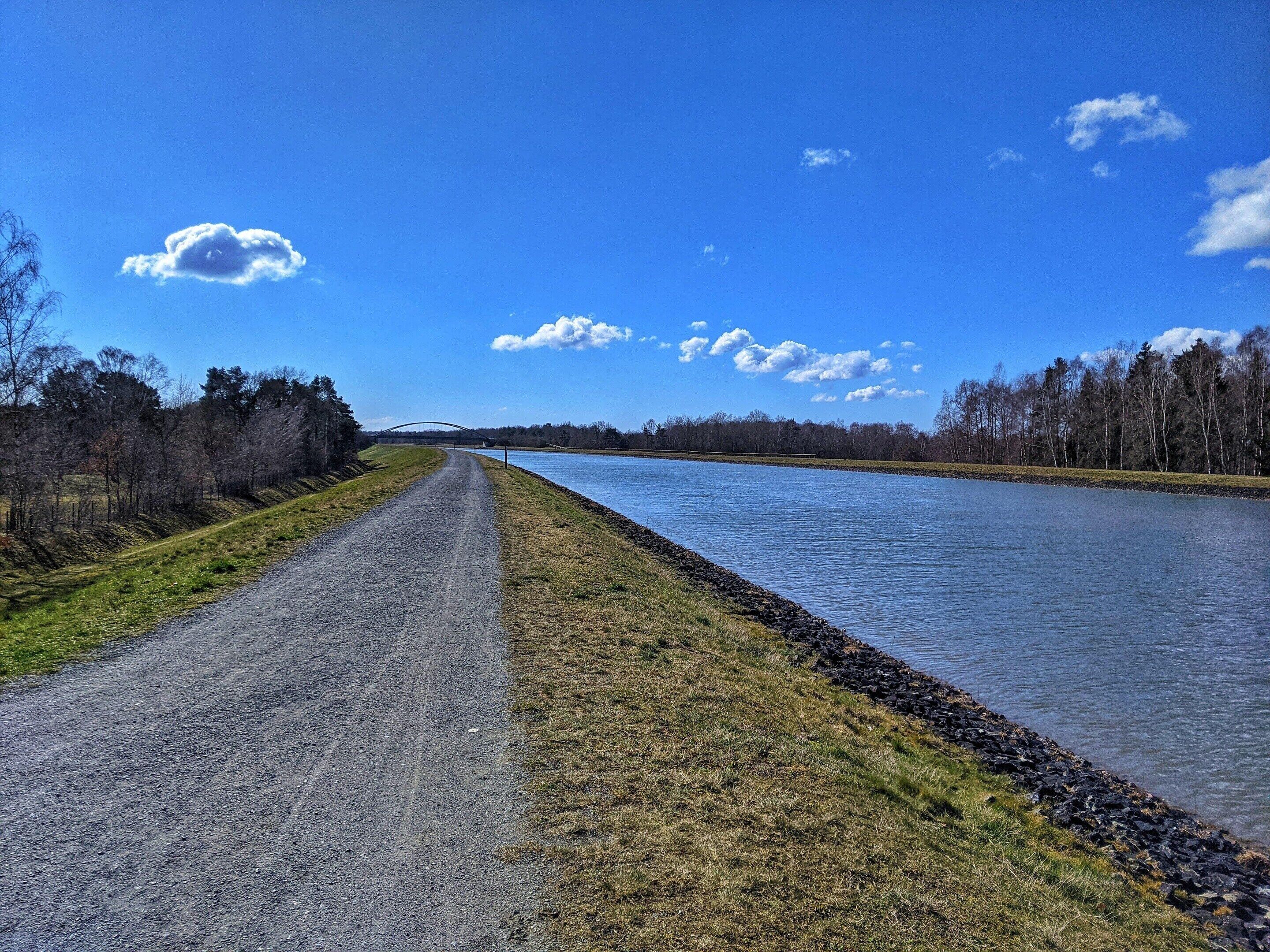 Un chemin de gravier longe une rivière, bordée d’arbres des deux côtés, sous un ciel bleu clair.