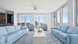 A spacious living room with two sofas, a coffee table, and a view of the city through large windows. - Luxury 4BR Condo w/ private beach access & heated pool (Gulf Shores)
