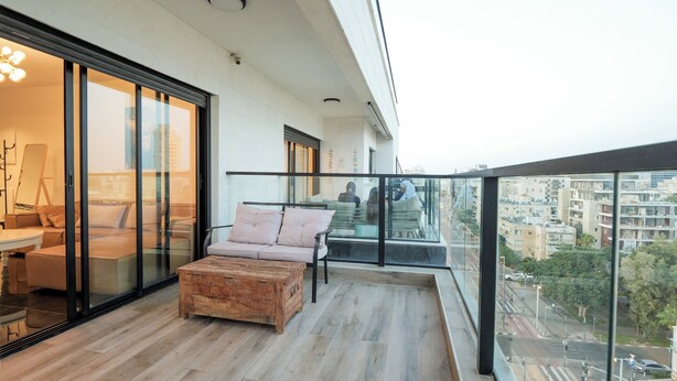 A modern balcony with a wooden bench, a cushioned chair, and a glass door leading to a room with a table and chairs. - Spacious 5-Room Apartment Near the Sea – Bat Yam (Bat Yam)