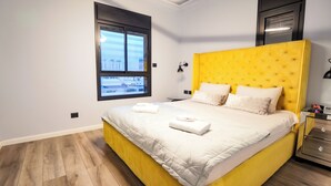 A modern bedroom with a yellow headboard, a large window, and a bedside table with a clock. - Spacious 5-Room Apartment Near the Sea – Bat Yam (Bat Yam)