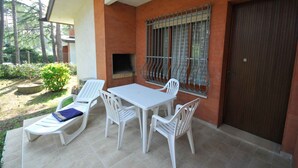 Un patio exterior con mesa y sillas blancas, una parrilla y vistas del follaje circundante.