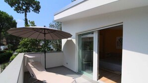 A modern balcony with a beige lounge chair and a large umbrella, a glass door leading to an interior room, and a clear view of the surrounding greenery. - Holiday apartment with air conditioning, terrace and garden (Lignano Sabbiadoro)