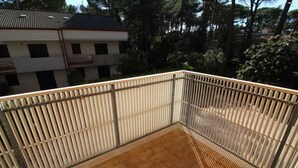 A balcony with metal railings, a tiled floor, and a view of a building with a tiled roof and trees in the background. - Holiday apartment with air conditioning and balcony (Lignano Sabbiadoro)