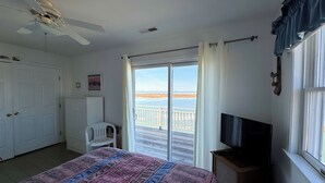 Ein Schlafzimmer mit Bett, Fernseher, Deckenventilator, Fenster mit Blick auf den Strand und einer Tür.