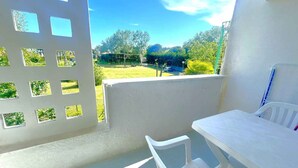Un balcon avec une table et une chaise blanches, une vue sur un paysage verdoyant et un mur blanc orné d’une grille de fenêtre.