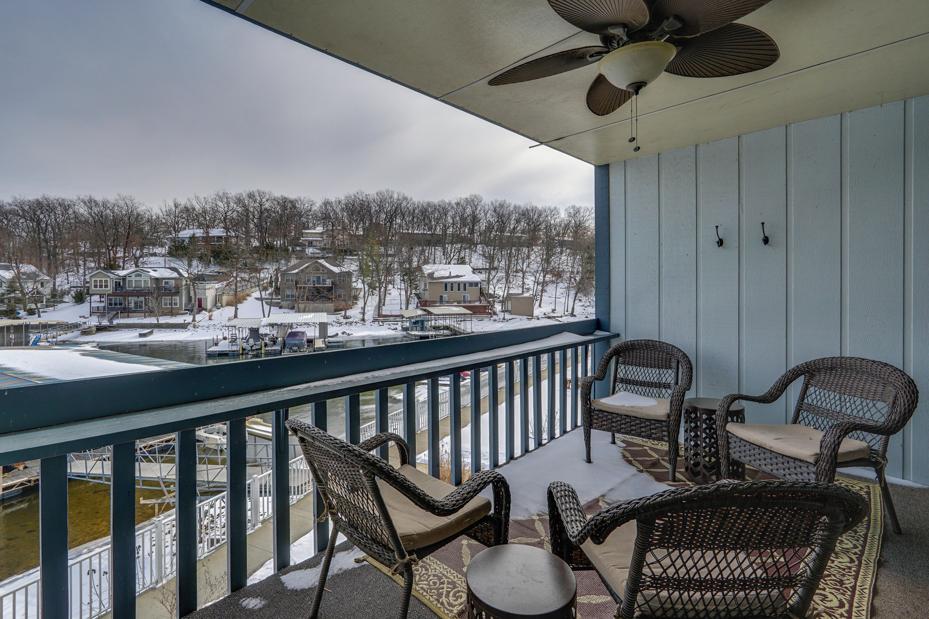 A balcony with wicker chairs and a table, overlooking a snowy residential area with houses and a dock.