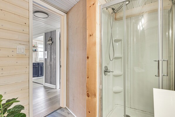 A modern bathroom with a glass shower enclosure, wooden paneling, and a visible kitchen area in the background.