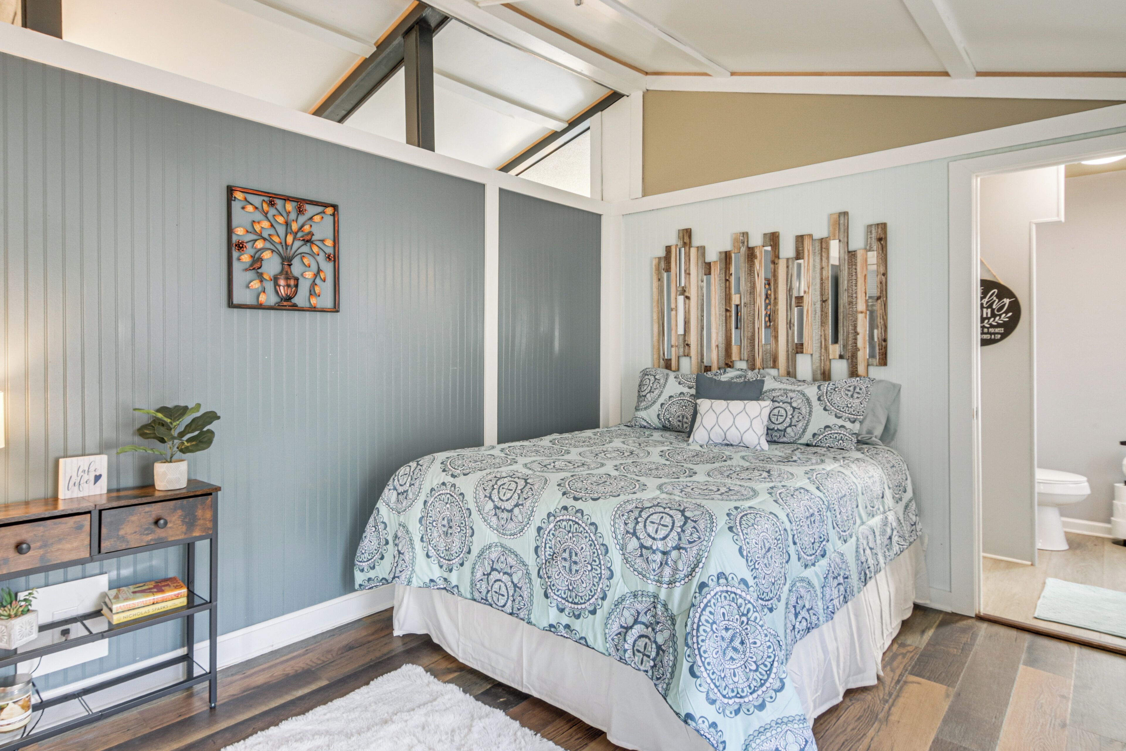 A bedroom with a bed, a nightstand, a potted plant, and a wall with a decorative piece.