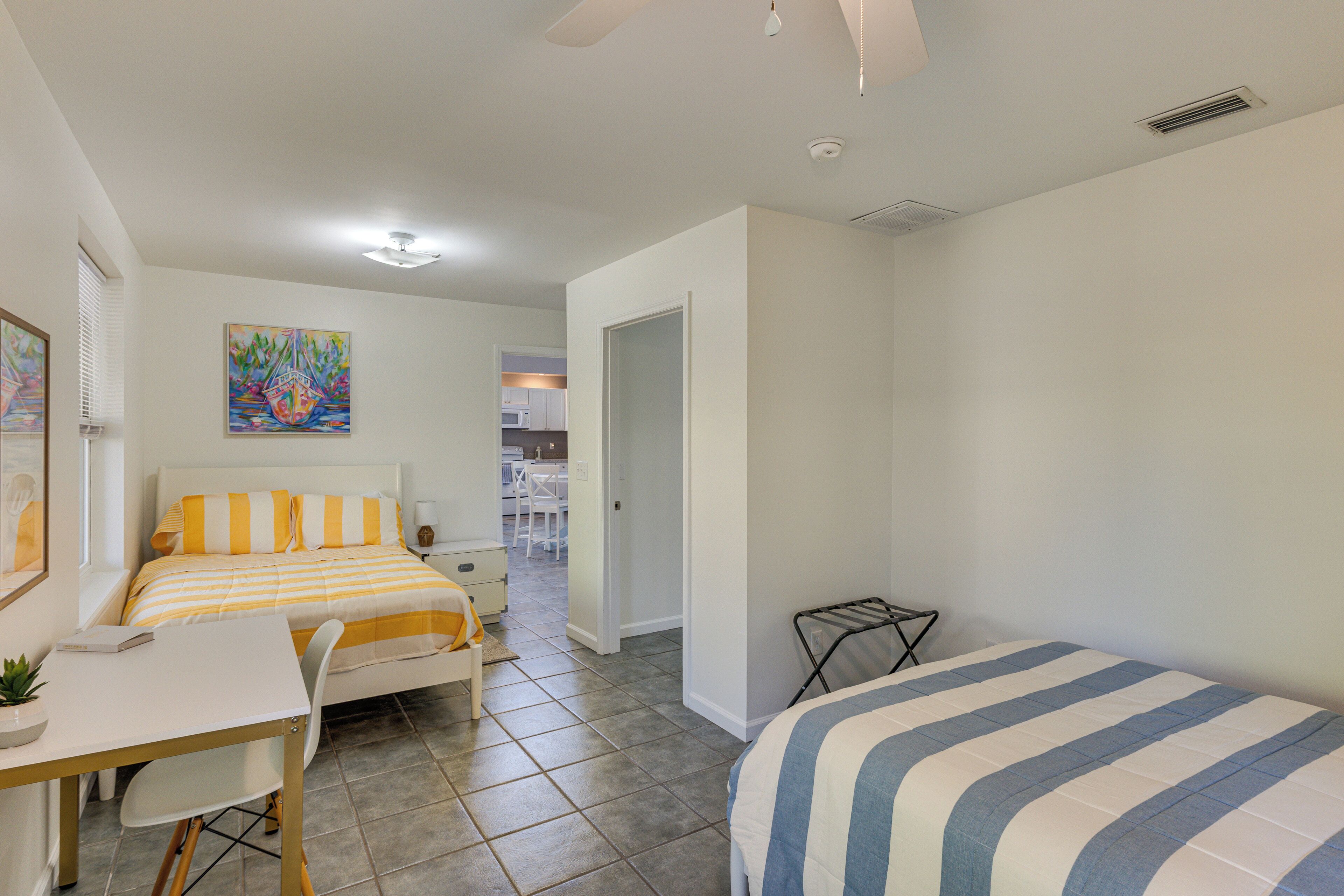 Un dormitorio con cama, escritorio, silla y una mesita. Hay un cuadro en la pared y una puerta que da a otra habitación.