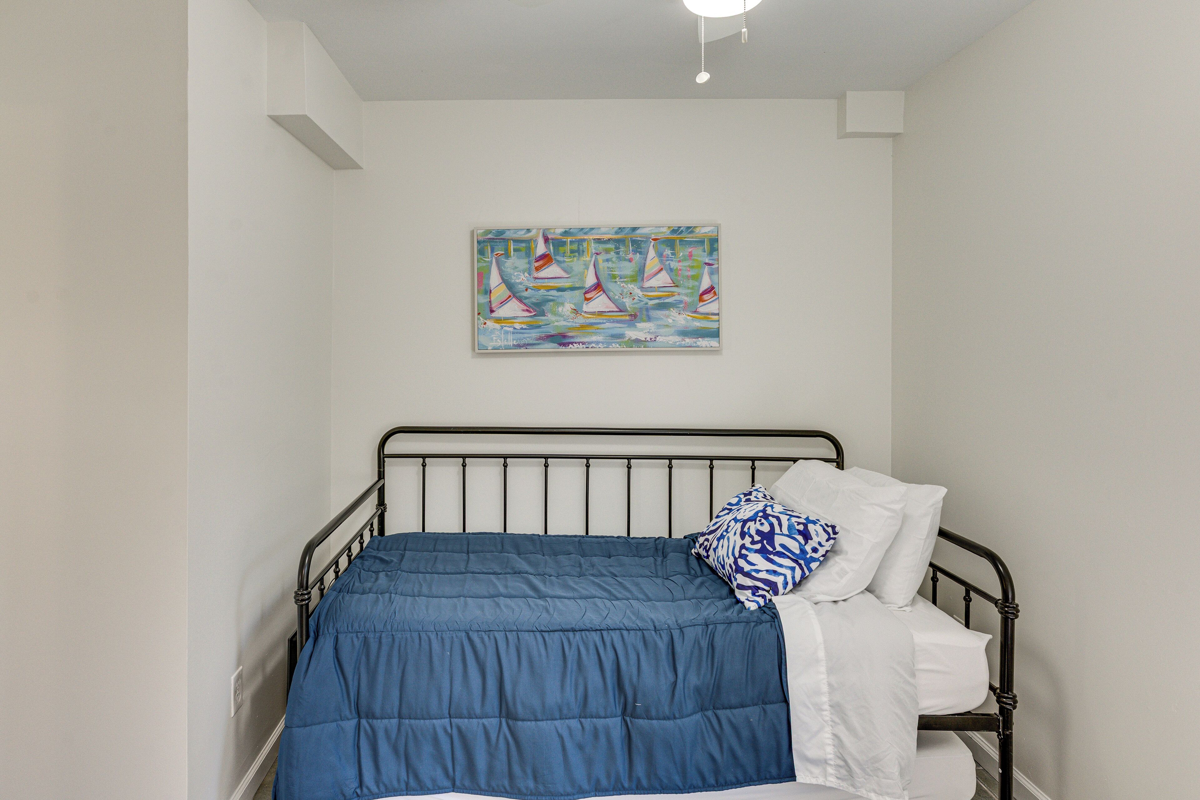 Een enkel bed met een blauwe deken, witte kussens en een schilderij van een zeilboot aan de muur.