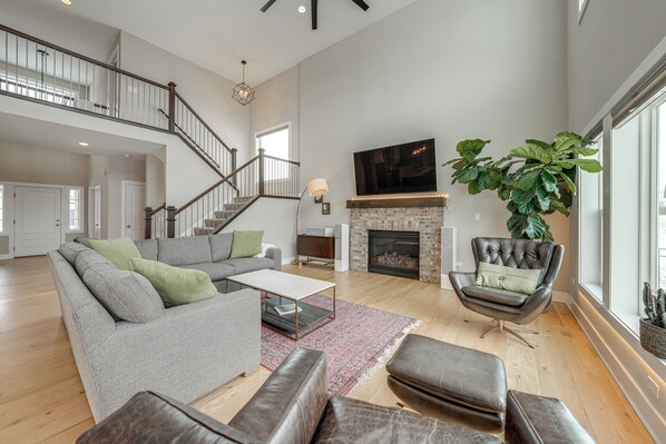 A spacious living room with a grey sofa, leather ottomans, a coffee table, and a fireplace. A large plant is by the window, and a staircase leads to an upper level. - Bring the Family! Suburban Gem in Plainfield (Plainfield)