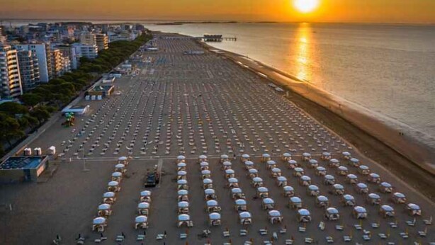 Uma praia com fileiras de guarda-sóis e cadeiras, um cais e edifícios ao fundo.