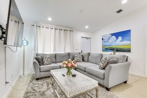 Uma sala de estar moderna com um sofá modular cinza, uma mesa de centro com um vaso de flores, uma televisão de tela plana e uma pintura de paisagem.