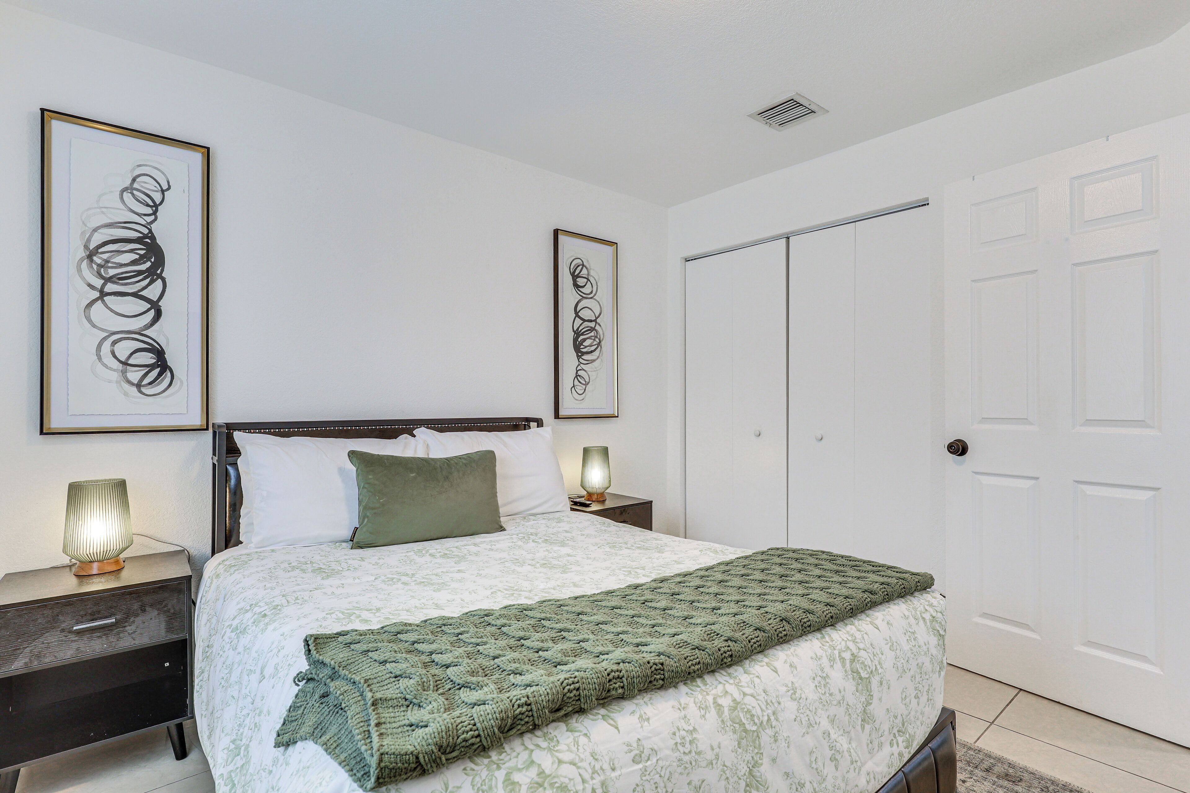 A bedroom with a bed, two bedside tables with lamps, a green throw blanket, and two framed abstract artworks on the walls.