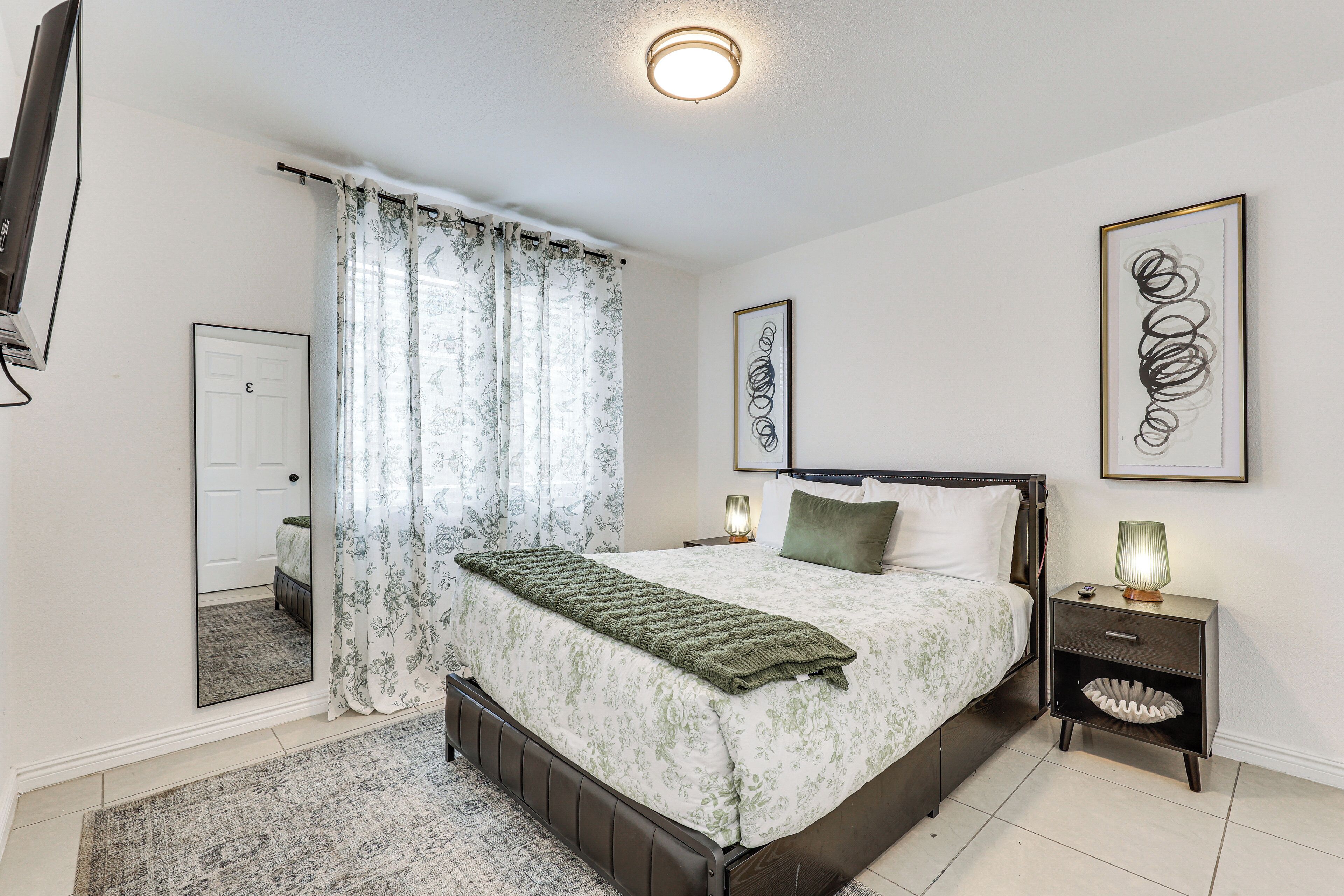 A bedroom with a bed, a nightstand, a mirror, a door, and a wall with framed artwork.