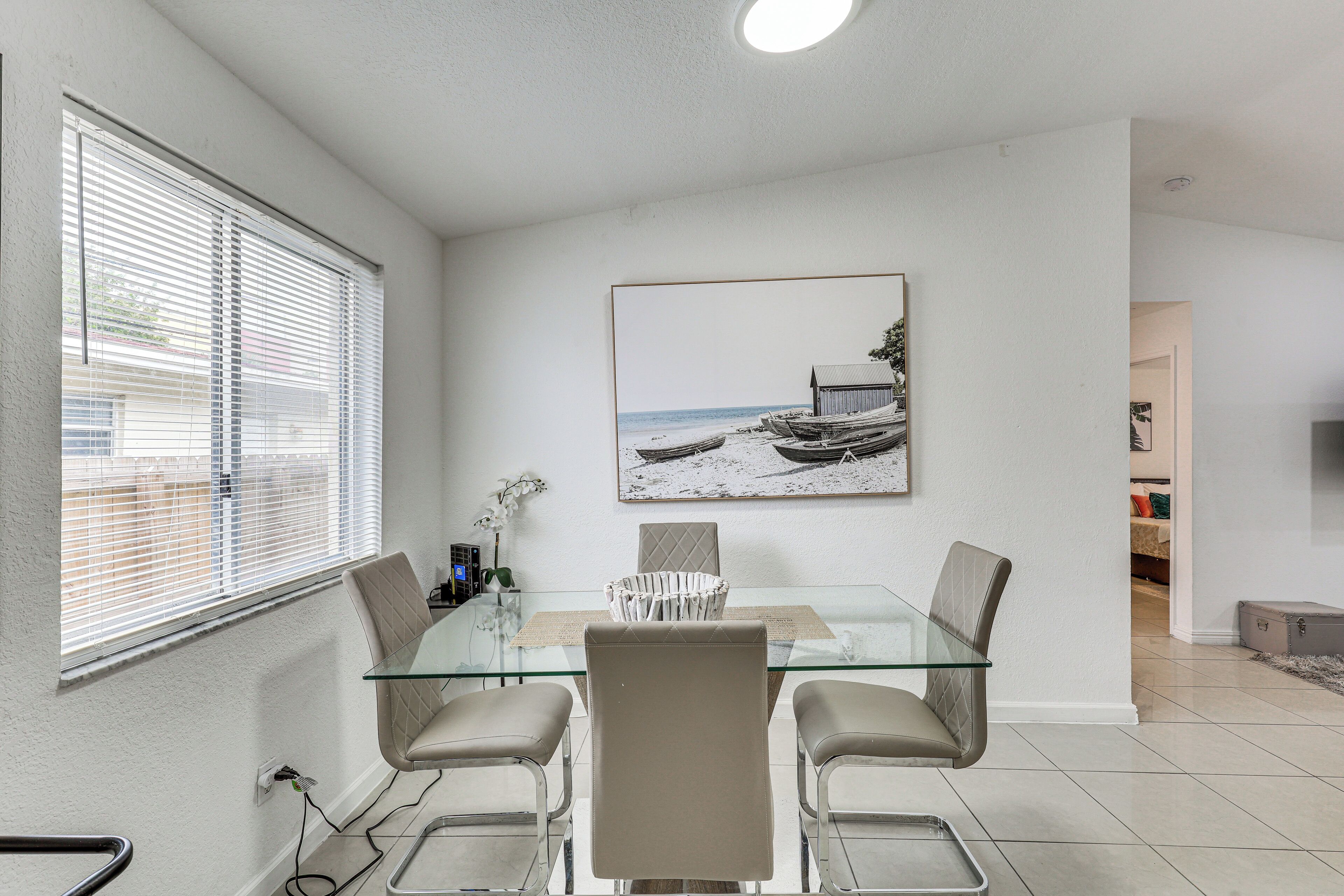 A dining area with a glass table, four chairs, a painting of a beach scene, and a window with blinds.