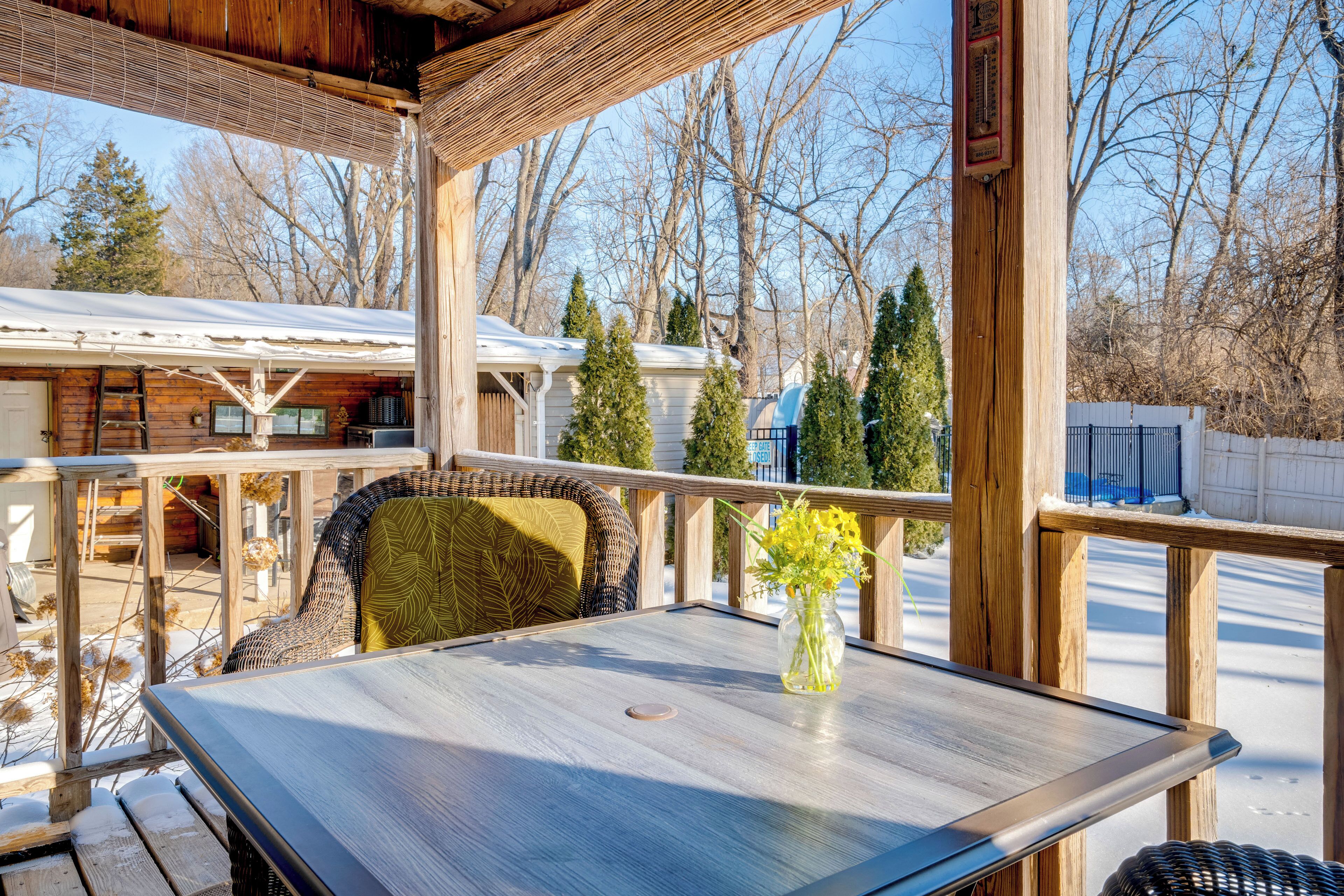 A wooden deck with a table and chairs, a vase with flowers, and a view of a cabin and trees.