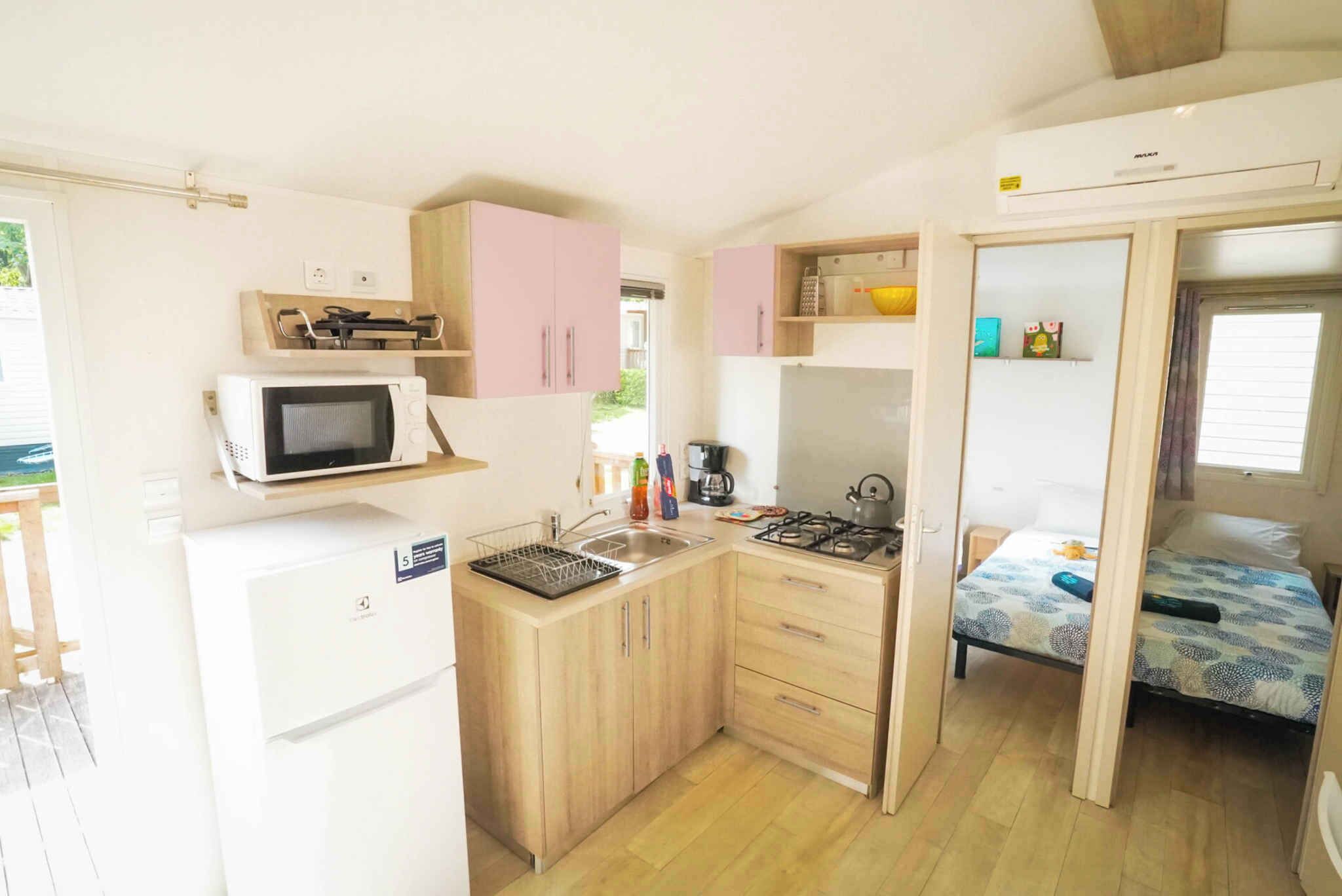 A compact kitchen with a microwave, sink, and stove, adjacent to a small bedroom with a bed and a window.