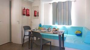 A small room with a blue sofa, a table with chairs, a window with striped curtains, and two fire extinguishers on the wall. - Mobile home with air conditioning, terrace and pool (Calella de la Costa)