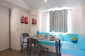 Ein kleiner Raum mit einem blauen Sofa, einem Tisch mit Stühlen, einem Fenster mit gestreiftem Vorhang und zwei an der Wand befestigten Feuerlöschern.