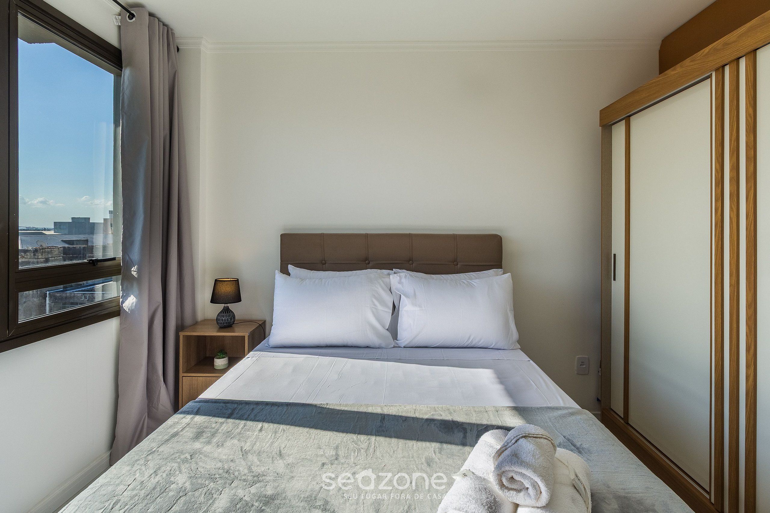 Ein Hotelzimmer mit Bett, Nachttisch, Lampe, Blick auf die Stadt und einem Kleiderschrank.
