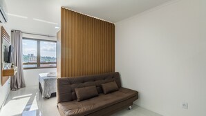A modern living room with a brown sofa, a wooden panel wall, and a window with a city view. - Full Studio 700m From Bourbon Shopping Umb0604 (Porto Alegre)