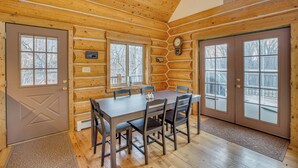 Una cabaña de troncos con comedor que incluye una mesa y sillas de madera, ventanas amplias y un reloj en la pared.