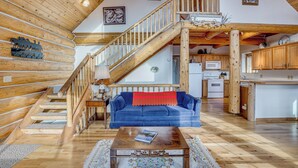 Interior de una cabaña de troncos con una escalera de madera, un sofá azul, una mesa de centro de madera y una zona de cocina.