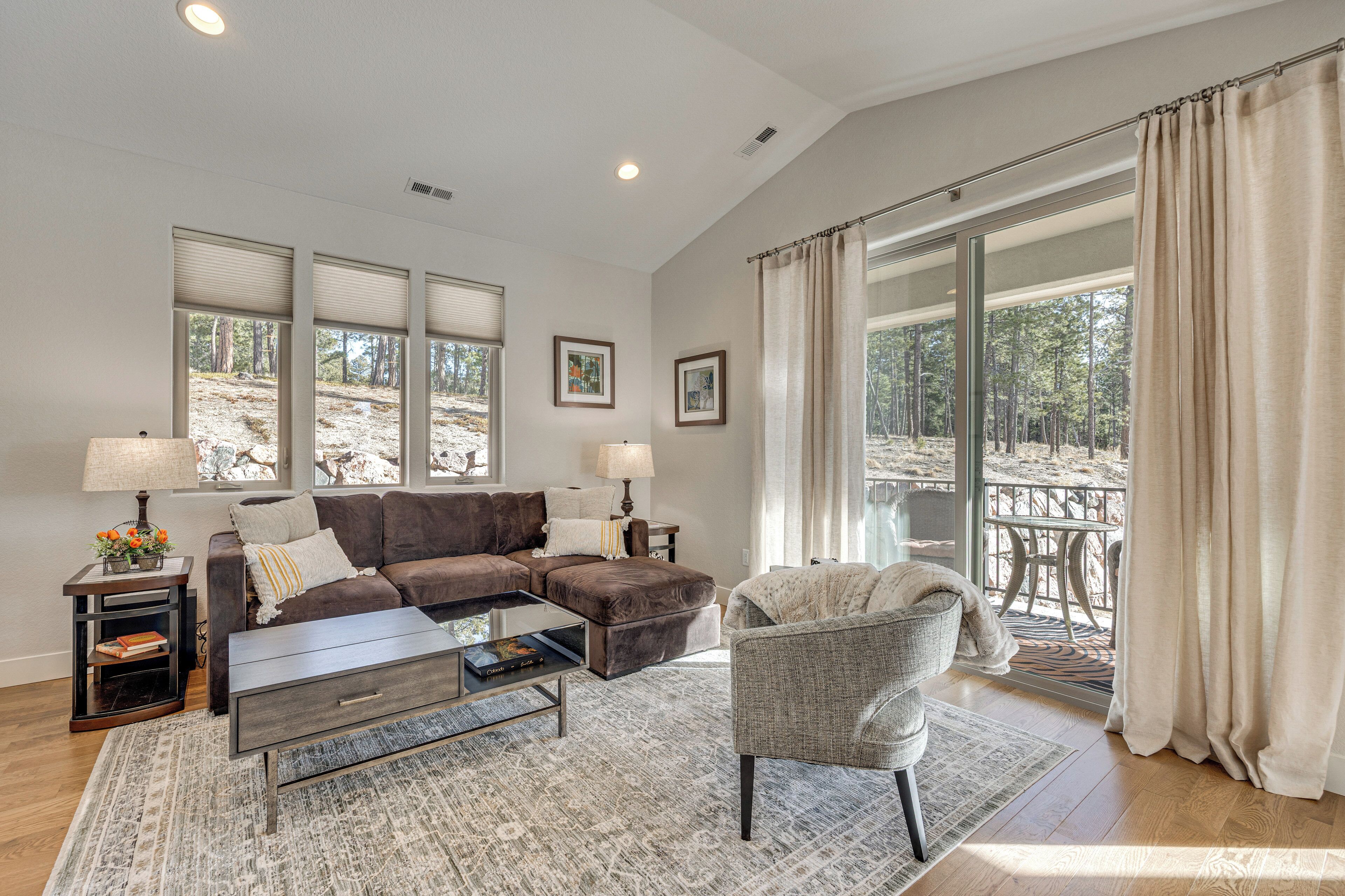 A spacious living room with a sectional sofa, a coffee table, and a chair. There are two side tables with lamps, a rug, and large windows with blinds.