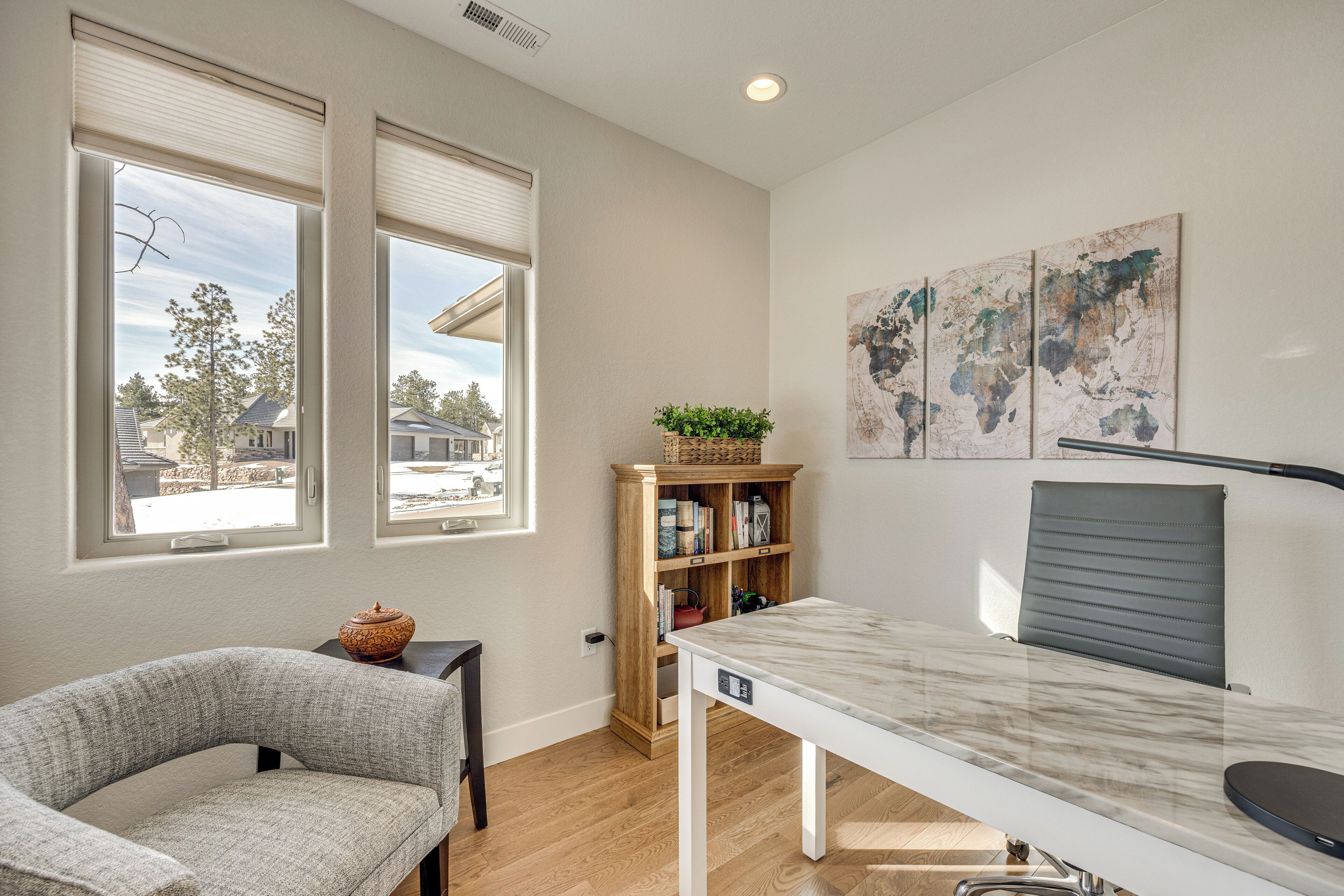A modern room with a curved grey chair, a white table with a grey chair, a wooden bookshelf with books and a plant, and a world map on the wall.
