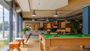 A billiards table with a green felt surface, a pool cue, and a cue ball. There are wooden stools and a small table with a potted plant. The room has large windows, a brick wall, and shelves with plants. - Cathedral Road by UTDM (Derby)