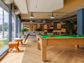 A billiards table with a green felt surface, a pool cue, and a cue ball. There are wooden stools and a small table with a potted plant. The room has large windows, a brick wall, and shelves with plants. - Cathedral Road by UTDM (Derby)