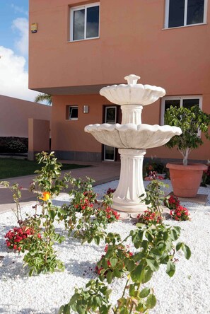 A white stone fountain with multiple tiers, surrounded by gravel and flowering plants, in front of a building with a brown facade and windows.