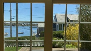Une vue depuis une pièce dotée d’une grande fenêtre donnant sur une terrasse avec des chaises blanches et un plancher en bois.