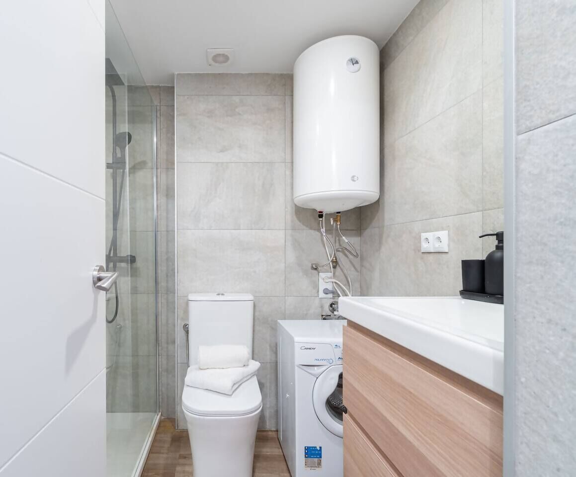 Een kleine badkamer met een douchecabine van glas, een toilet, een wasmachine en een wit wastafelmeubel met een zwarte zeepdispenser.