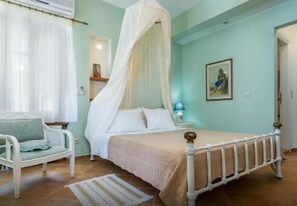 A bedroom with a four-poster bed, a bedside table with a lamp, a chair, and a painting on the wall. - Stolios Skopelos - Clio Superior Panoramic Seaview (Skopelos)
