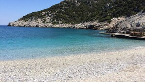 Ein Kiesstrand mit klarem türkisblauem Wasser und einer felsigen Hügelkulisse im Hintergrund.