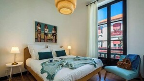 Une chambre à coucher avec un lit, deux lampes de chevet, un tableau, une fenêtre donnant sur un beau paysage et un fauteuil.