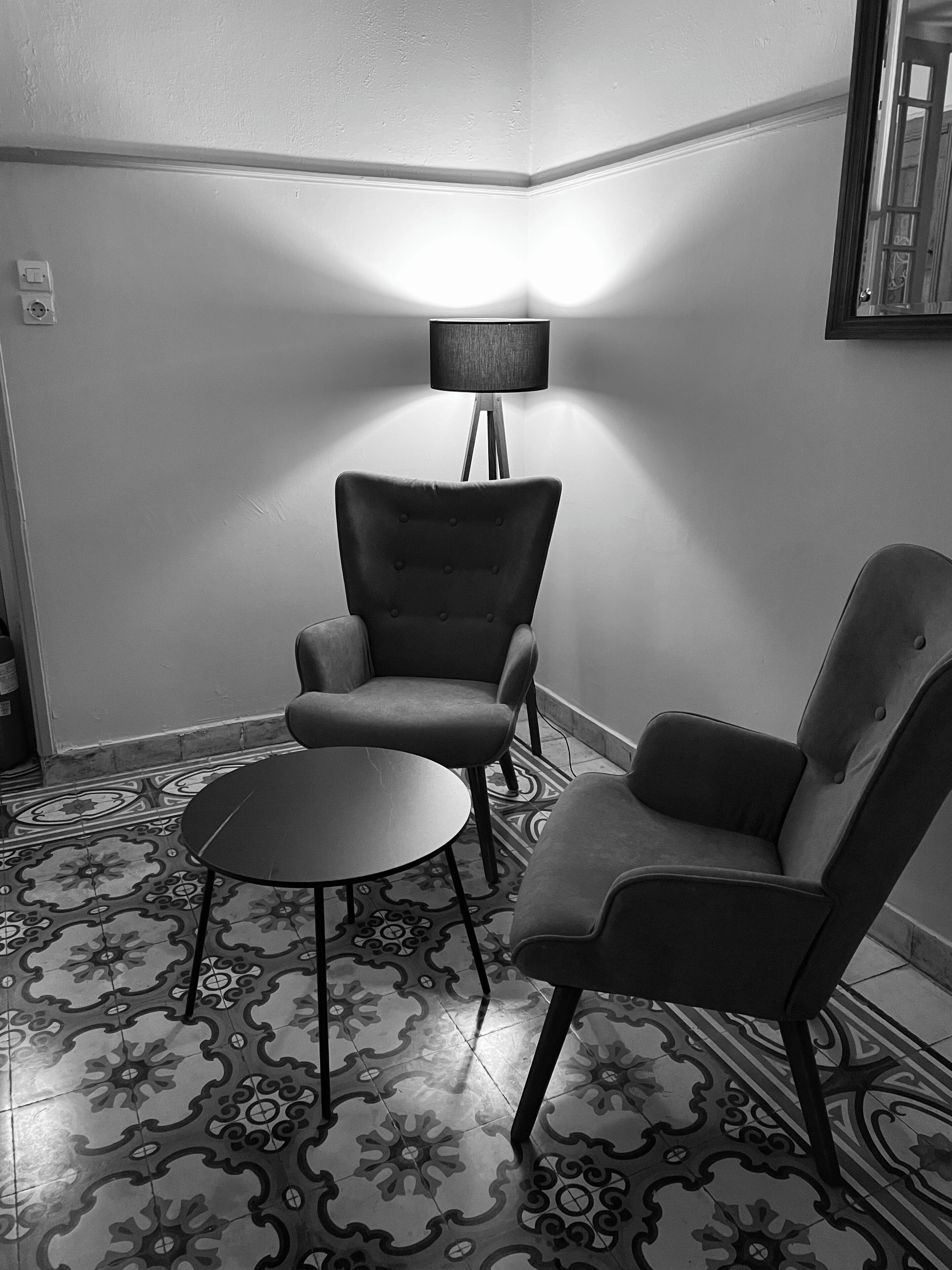 A corner room with two armchairs and a small round table. A floor lamp is positioned between the chairs. The floor has a patterned tile design.